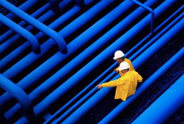 Two oil workers stand beside a network of pipes.