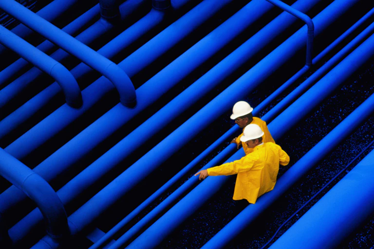 Two oil workers stand beside a network of pipes.