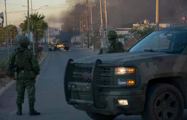 Two soldiers wearing camouflage stand in front of a dirty SUV and look at a burning vehicle in the middle of a street, with smoke going up in the air.