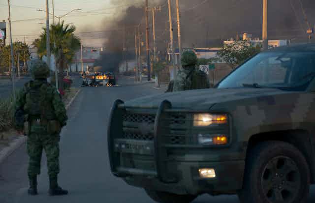 Two soldiers wearing camouflage stand in front of a dirty SUV and look at a burning vehicle in the middle of a street, with smoke going up in the air. 