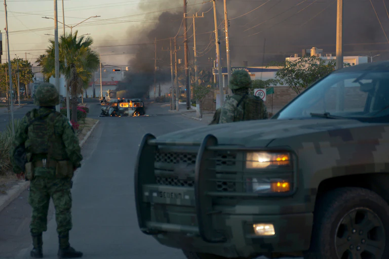 Two soldiers wearing camouflage stand in front of a dirty SUV and look at a burning vehicle in the middle of a street, with smoke going up in the air.