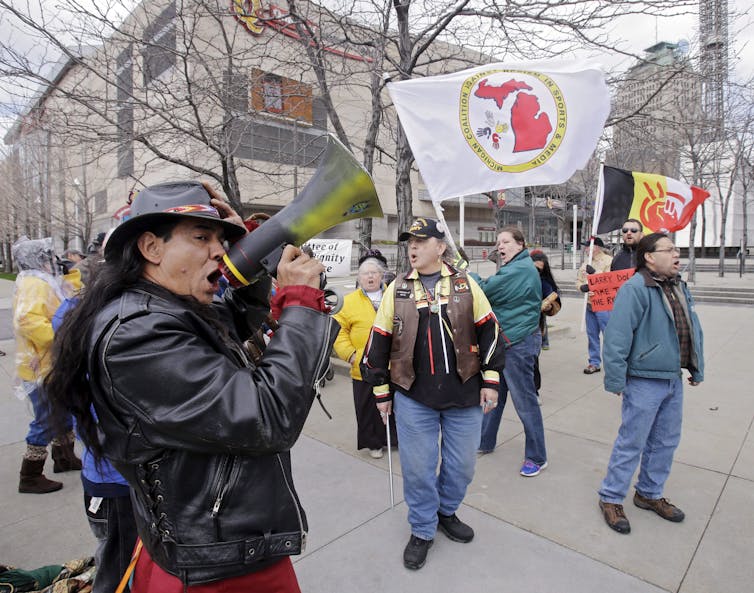 A man with long, dark hair speaks into a megaphone while attending a protest