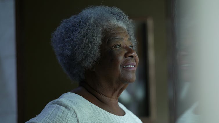 An elderly black woman with grey hair looking out of a window.