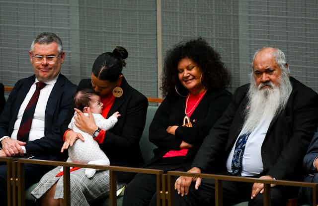 Four people sit in Parliament. Senator Jana Stewart is sitting with her baby, alongside Patrick Dodson and Malarndirri McCarthy.
