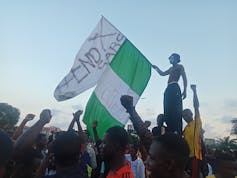 Man standing on a platform and waving a flag and surrounded by people raising their hands in protest.