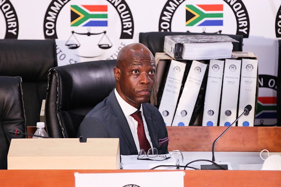  A seated man wearing a suit and tie stares ahead. Behind him is the logo of South Africa's state capture commission.