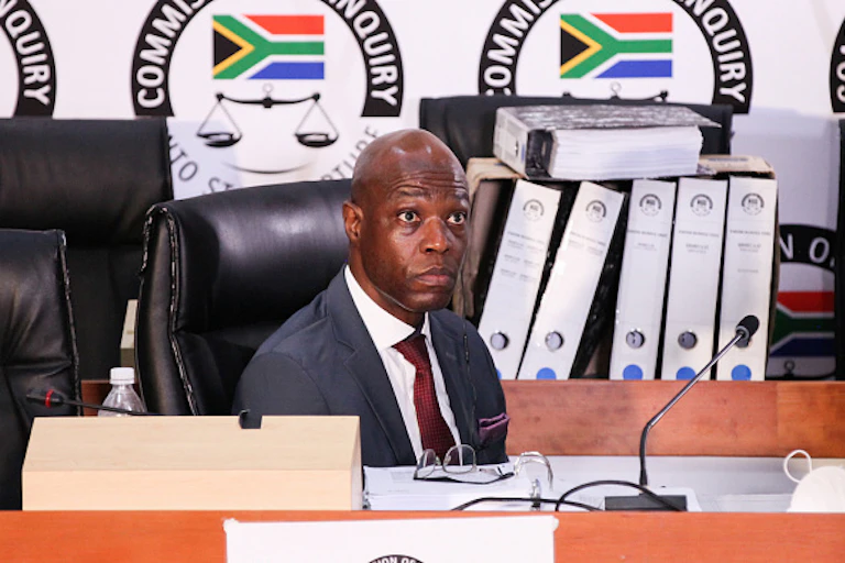 A seated man wearing a suit and tie stares ahead. Behind him is the logo of South Africa's state capture commission.