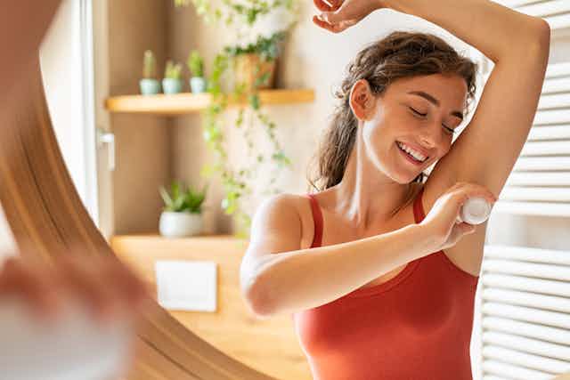 Un espejo refleja a una mujer sonriente con camiseta de tirantes roja mientras se aplica desodorante en el sobaco.