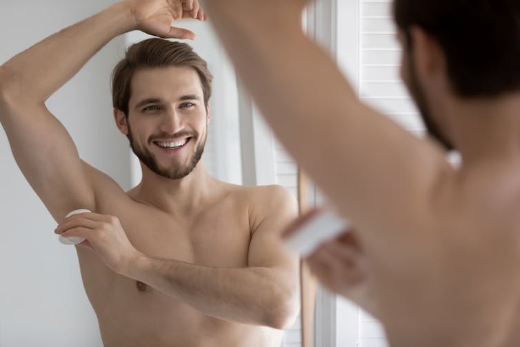 Un hombre con barba sonriente con el torso desnudo sonríe a un espejo mientras se aplica desodorante en el sobaco.