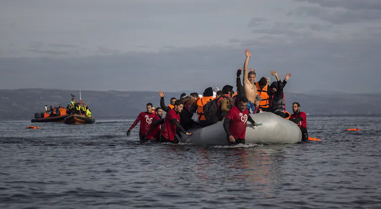 A bare-chested man raises his hands in triumph at the front of a dinghy filled with people wearing life jackets.