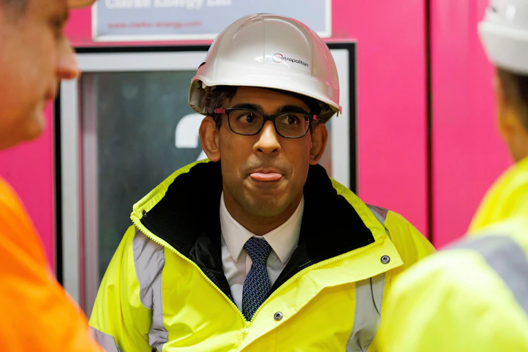 Rishi Sunak wearing a helmet and high viz jacket, sticking out his tongue.