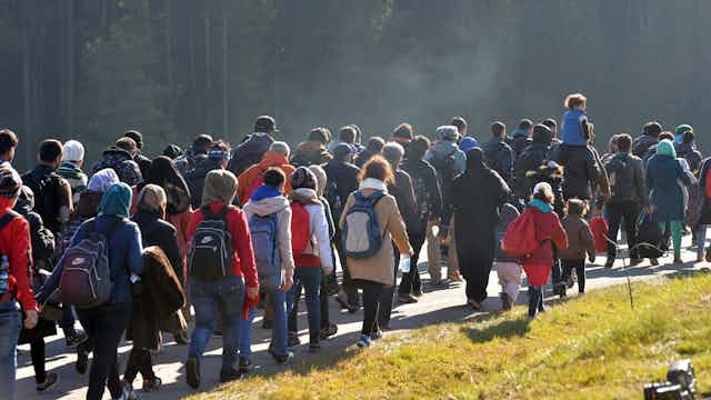 A large group of people carrying knapsacks and other belongings walk along a road.