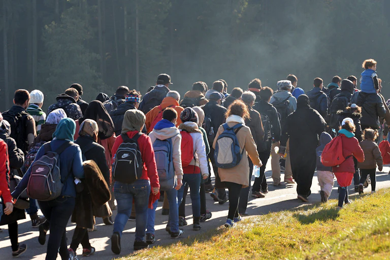 A large group of people carrying knapsacks and other belongings walk along a road.
