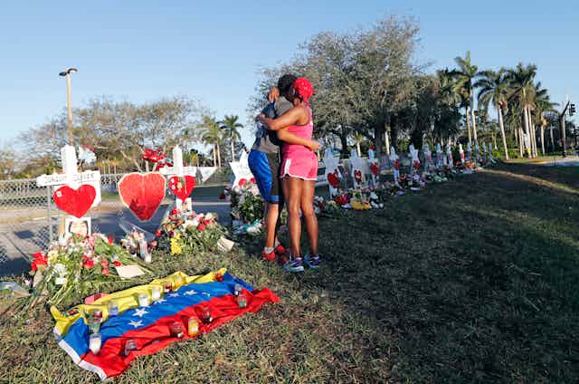 Two people hug in front of a  line of white crosses adorned with flowers