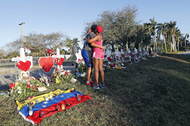 Two people hug in front of a line of white crosses adorned with flowers