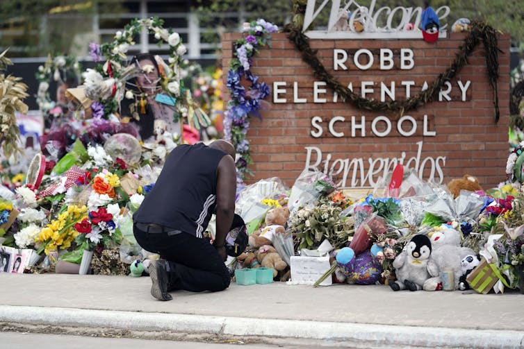 A man kneels in front of a brick wall saying 'Robb Elementary School,' with piles of flowers all around.