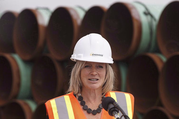A blonde woman in a white hard hat stands in front of large oil transportation pipes.