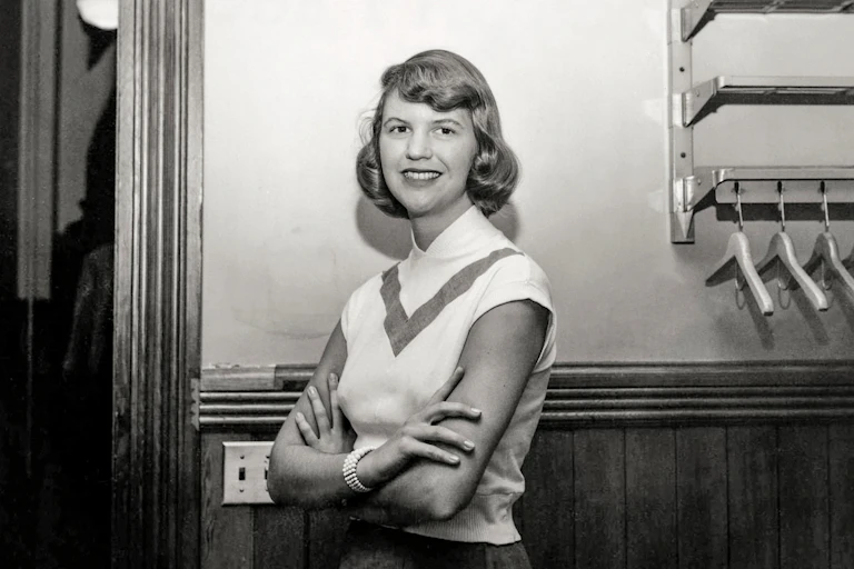 Sylvia Plath poses with her arms crossed, smiling.