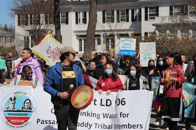 A man with a drum, several people holding a banner and dozens of others behind them march in front of a large white home.