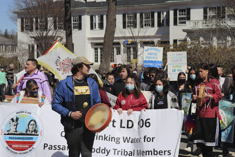 A man with a drum, several people holding a banner and dozens of others behind them march in front of a large white home.