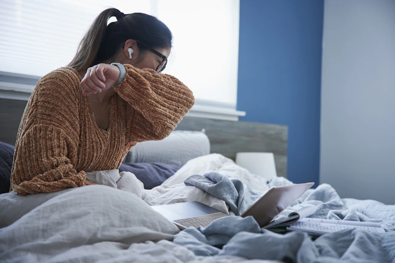 A woman coughing into her elbow while sitting in bed.