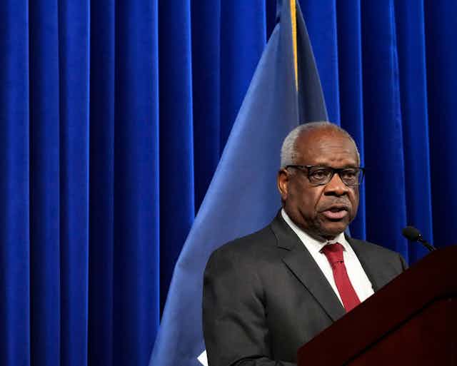 With an American flag in the background, a Black man wearing eyeglasses and dressed in a business suit is standing at a podium.