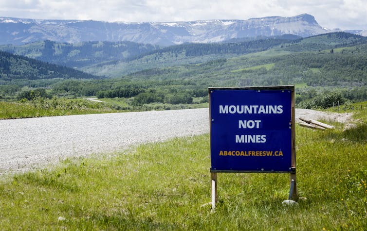 A sign says Mountains not Mines next to a road leading to a mountain range.