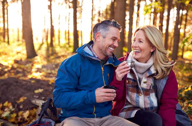 A man and woman, both smiling and with a hot drink in hand, stop for a rest during a walk in the country.