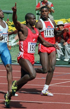 A man on a running track wearing a red vest and shorts raises his arm.