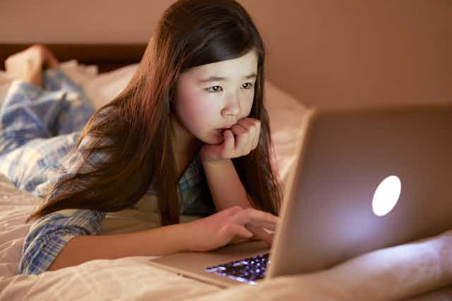a girl in pajamas lying on top of her bed using a laptop computer