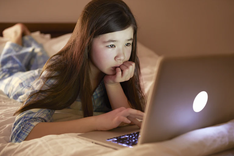 a girl in pajamas lying on top of her bed using a laptop computer