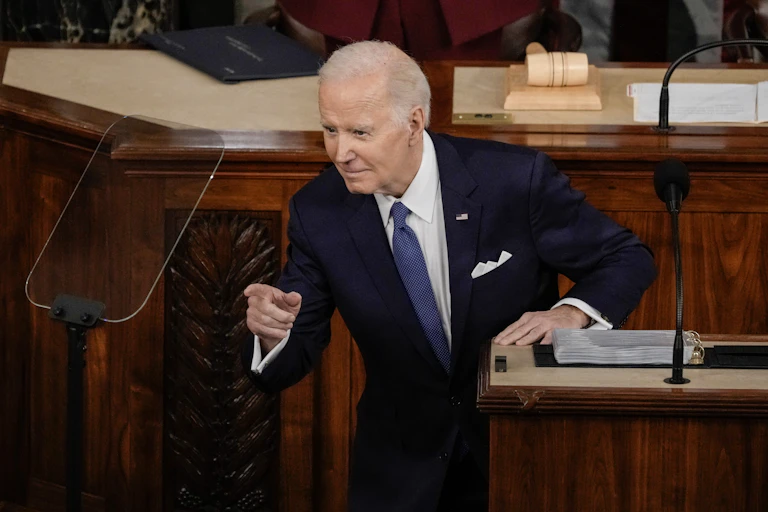 A gray haired man in a blue suit, white shirt and blue tie points his finger at someone not in the photo.