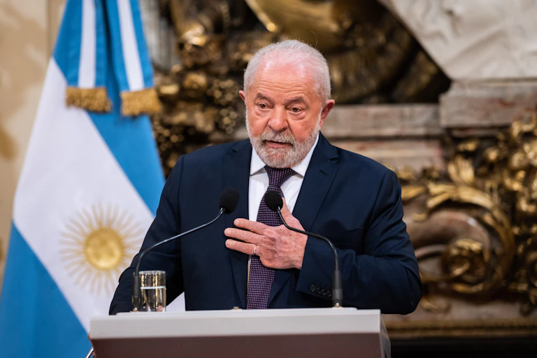 A gray-haired, suited man stands behind a lectern, speaking into a microphone as his three-fingered hand rests on his chest. Behind him, to his right stands a light blue flag with a broad white swath and and gold sun.