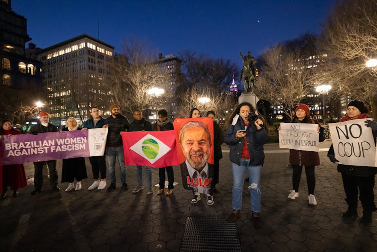 Un grupo de hombres y mujeres, con abrigos y sombreros de invierno, se paran al aire libre en una línea horizontal con carteles y pancartas.  La pancarta en el centro es la cara de un hombre canoso.  Debajo de la imagen aparece el nombre Lula.