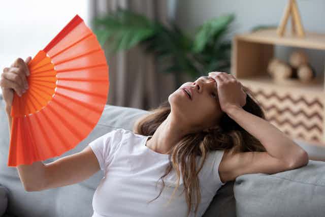 Woman on sofa holding forehead with one hand, waving fan at face with other