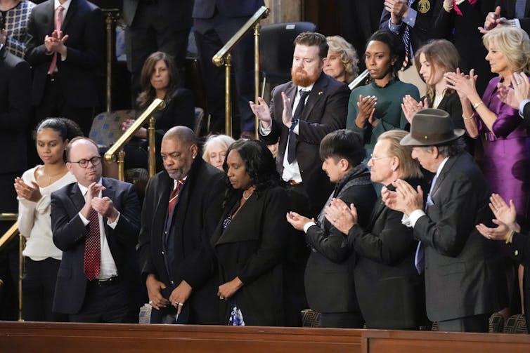 Two Black people stand with heads bowed as other people around them turn to face them and applaud.