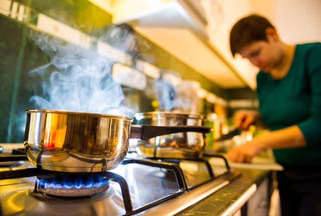 A person chopping ingredients in the background with a pan boiling on a gas stove in the foreground.
