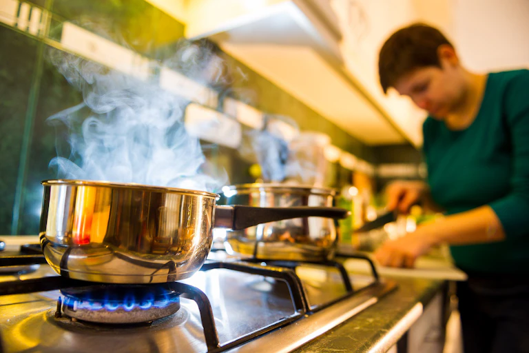 A person chopping ingredients in the background with a pan boiling on a gas stove in the foreground.