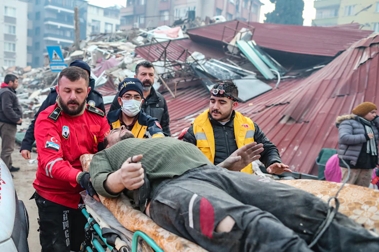 A man wearing a khaki top lies on a gurney. He is being pushed by a man in red and two people in yellow. Behind is a heap of rubble.