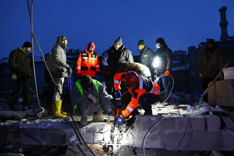 It is dark and a team of people use pneumatic equipment to cut through a block of concrete. They are operating under torchlight.