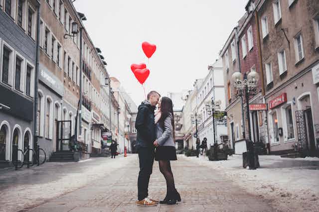 A man and woman embrace on a street while holding red heart-shaped balloons.