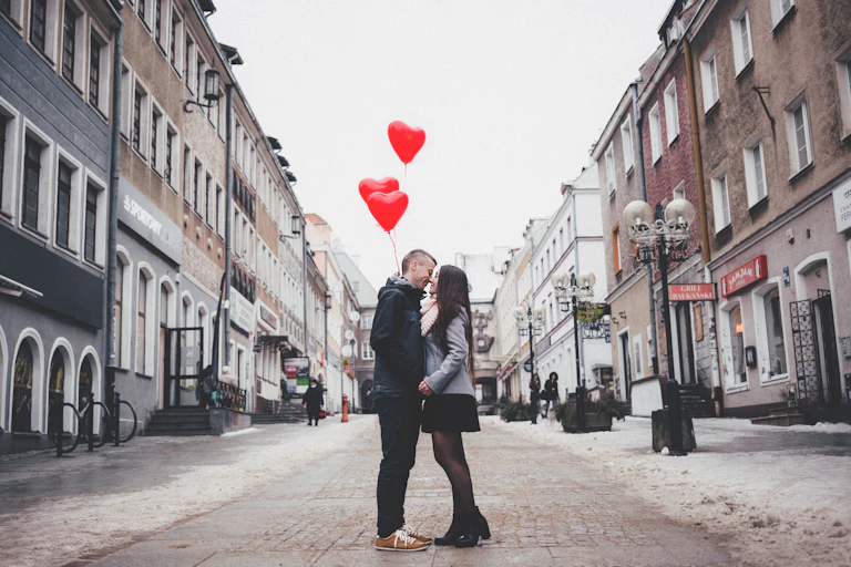 A man and woman embrace on a street while holding red heart-shaped balloons.