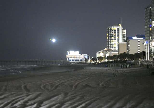 A dark photo shows a bright white circle over a dark beach, next to a lit up Ferris wheel and some buildings along the shore.