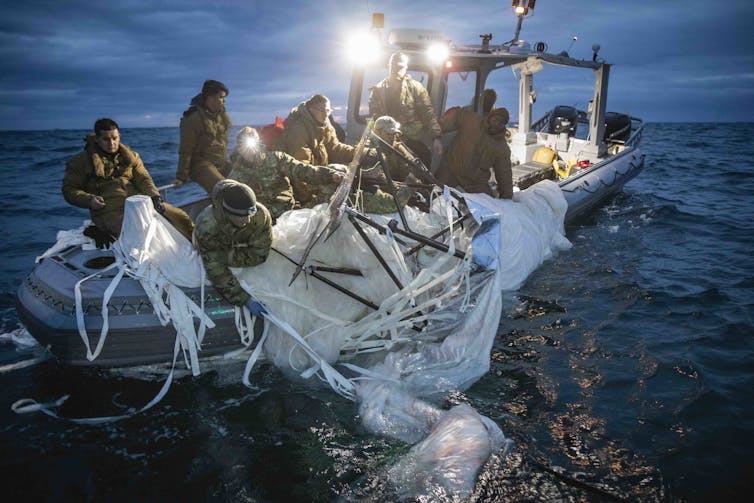 About 10 people wearing heavy clothing and camo sit on a small boat and lean over a large white plastic sheet, which has black sticks and white strings throughout.