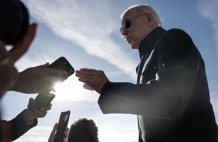 An older white man stands against a bright blue sky and talks to people, whose hands and voice recorders or phones are only visible.