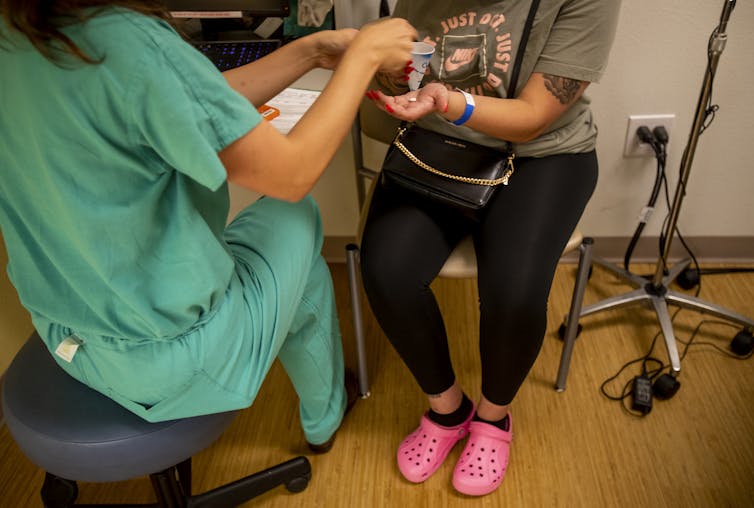 A woman in scrubs gives a pill to a woman in black leggings and pink Crocs