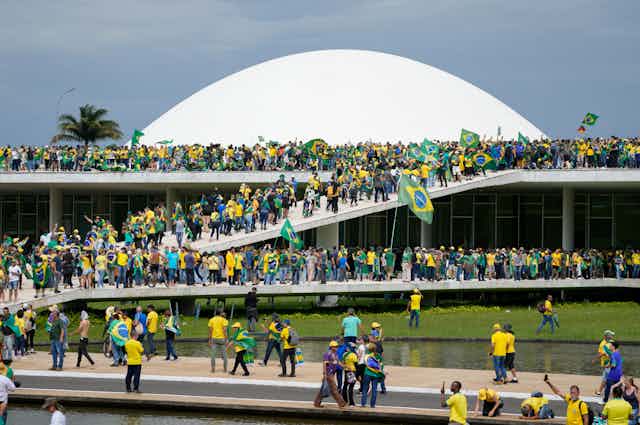 Hundreds of people storm a large building with a white dome, many carrying the Brazilian flag.