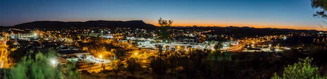 A large landscape shot of Alice Springs at sunset. Town shines with lights.