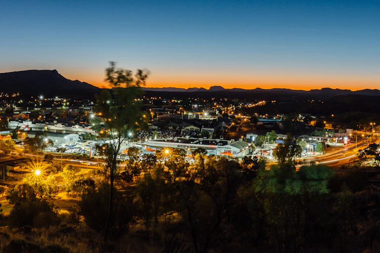 A large landscape shot of Alice Springs at sunset. Town shines with lights.