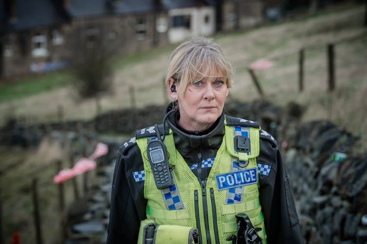 Sarah Lancashire looks to camera in her police uniform, with fields behind her.